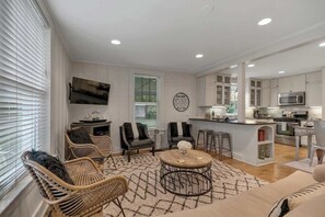 A modern living room with wicker furniture, a round coffee table, and a kitchen area with white cabinets and stainless steel appliances.