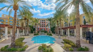 A pool area with palm trees, a red building in the background, and a clear blue sky.