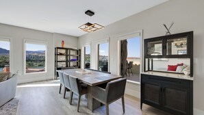 A modern dining area with a wooden table and chairs, a black cabinet with glass doors, and large windows offering a view of the city.
