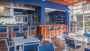 Un área de comedor al aire libre con sillas y mesas azules, una barra con mostrador de madera y una cocina con electrodomésticos de acero inoxidable.