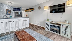 A modern kitchen with white bar stools, a wooden bench, a TV, and a fireplace. - Coastal Cozy Condo: Pet Friendly! Steps to Beach, Perfect for Relaxing Getaways! (Carolina Beach)