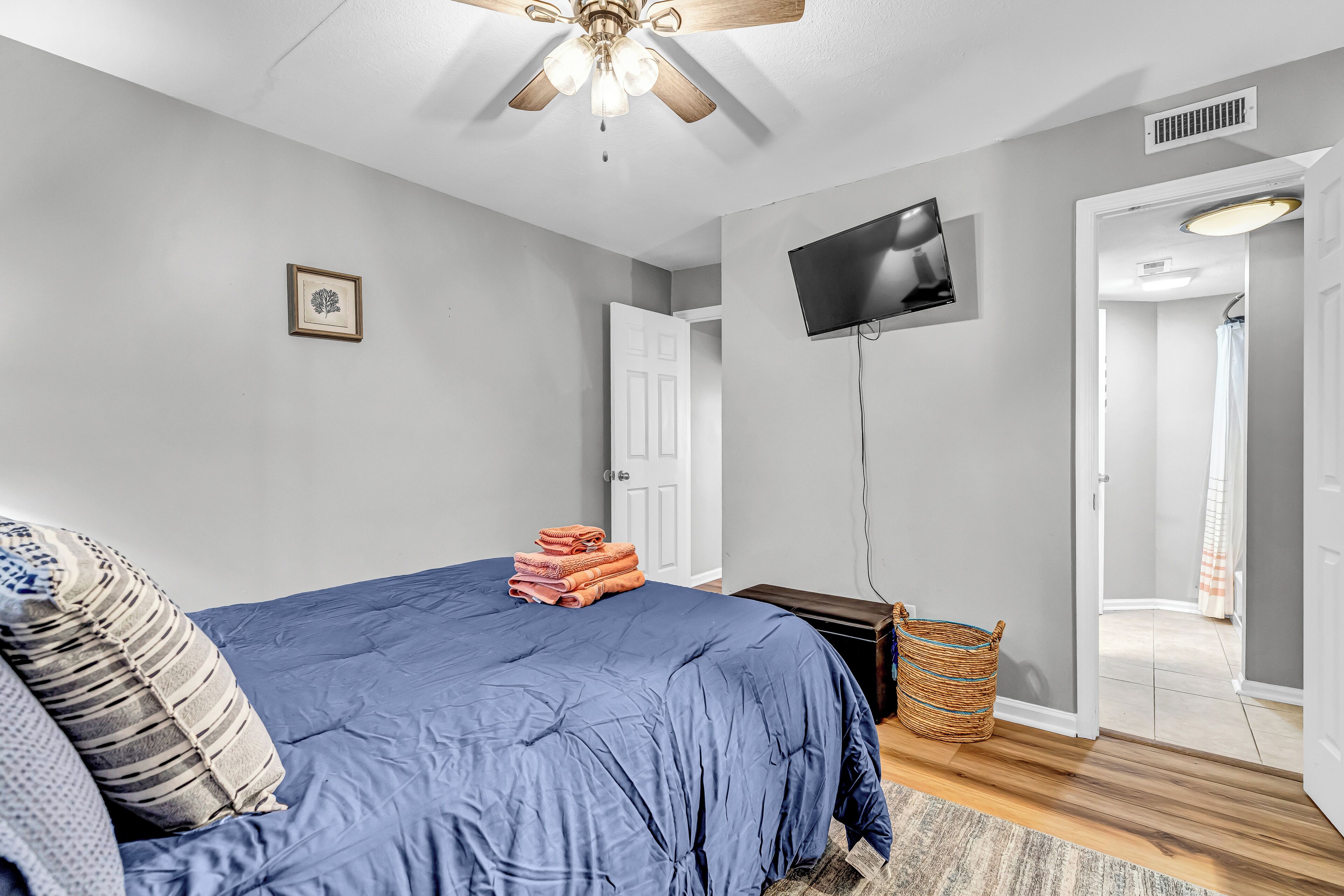 Un dormitorio con cama, ventilador de techo, televisor, un cuadro enmarco y una puerta que da a otra habitación.