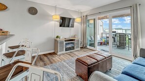 A modern living room with a dining area, a television, and a large sliding glass door leading to an outdoor patio. - Coastal Cozy Condo: Pet Friendly! Steps to Beach, Perfect for Relaxing Getaways! (Carolina Beach)
