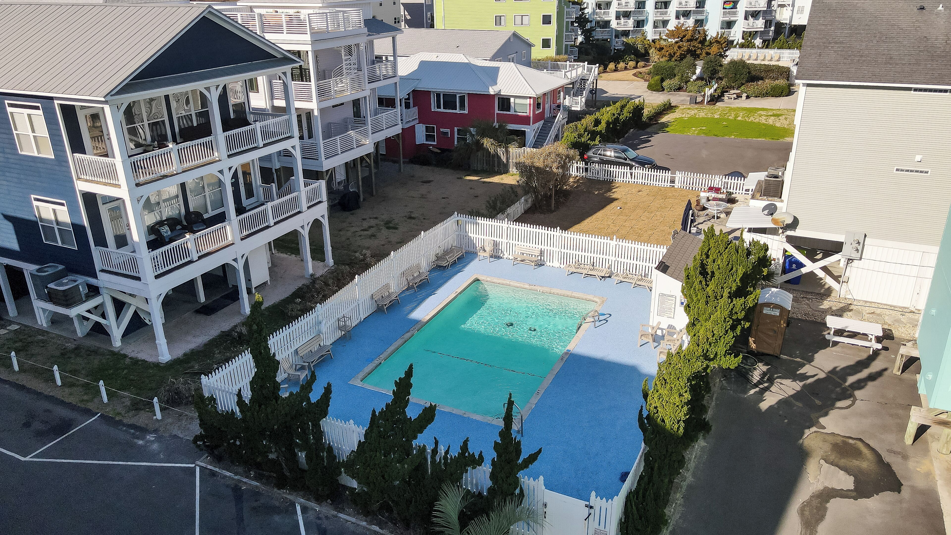 Ett bostadsområde med en swimmingpool omgiven av ett vitt staket, ett hus med flera våningar och balkonger samt en liten uteplats.