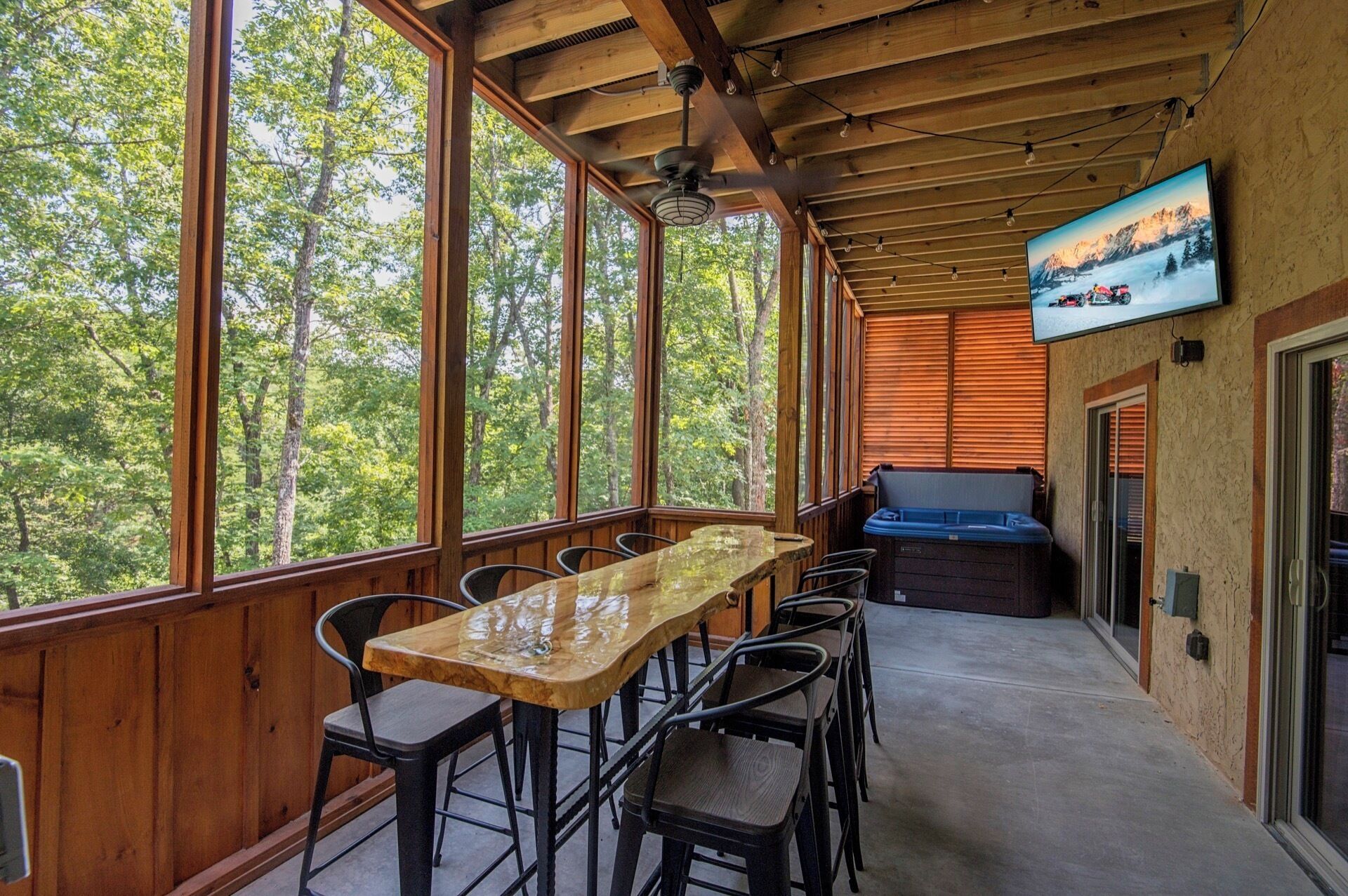 Un espace extérieur abrité, doté d’une table et de chaises en bois, d’un jacuzzi et d’un écran de télévision diffusant un paysage de montagne.