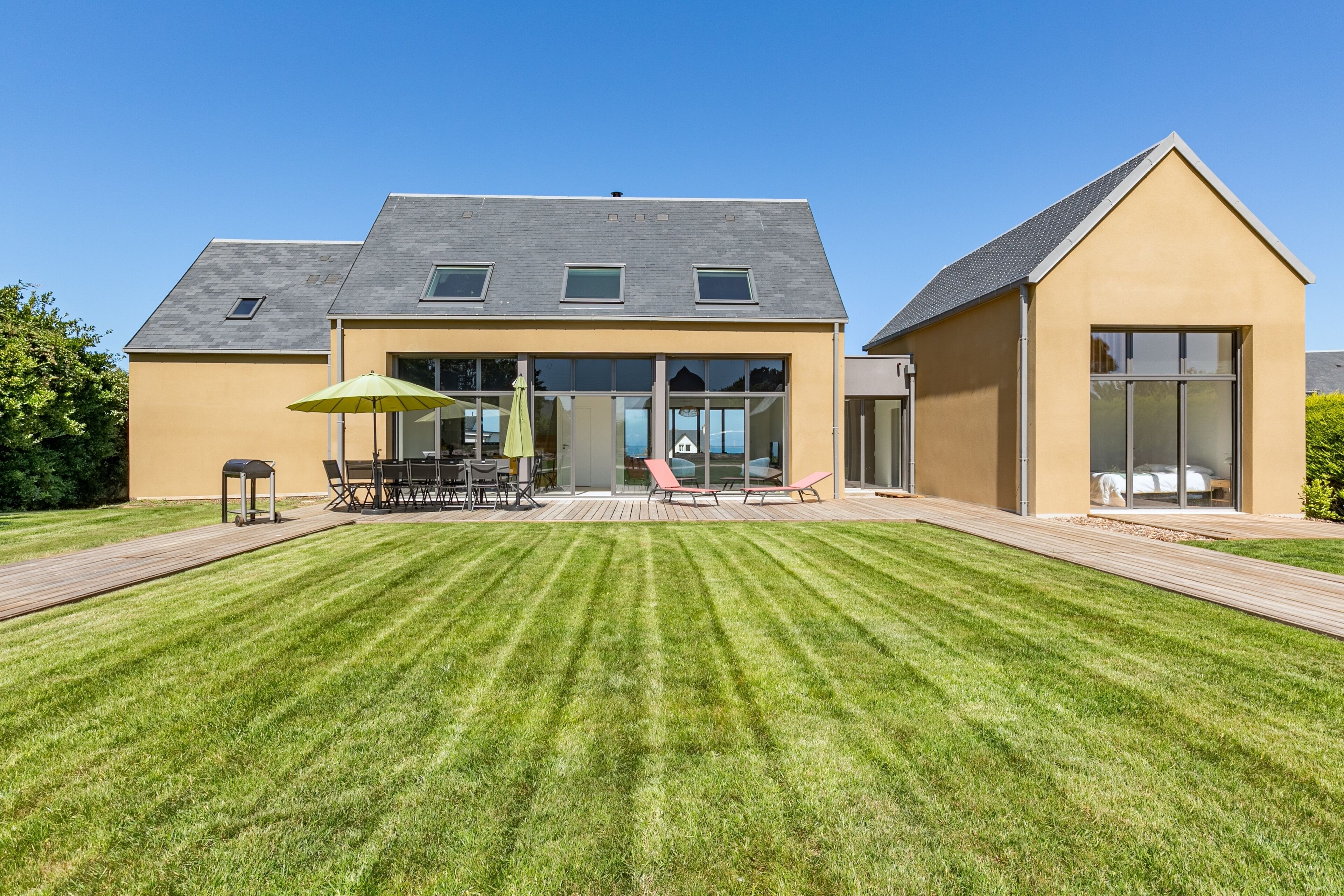 A modern house with a well-maintained lawn, a patio with outdoor furniture, and a large windowed room.