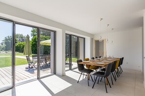 Un vaste espace dédié à la salle à manger, avec une grande table dressée pour six personnes, de grandes portes-fenêtres coulissantes donnant sur une terrasse et une vue sur l’extérieur.