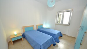 A bedroom with two beds, a nightstand with a lamp, a window with shutters, and a ceiling fan.