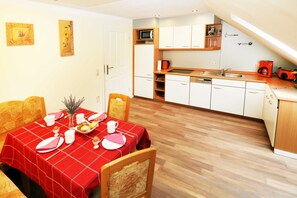 A dining area with a red tablecloth, wooden chairs, and a kitchen with white cabinets and a microwave. - Wolfgram Apartment with Balcony - Ferienwohnung Wolfgram (Trassenheide)