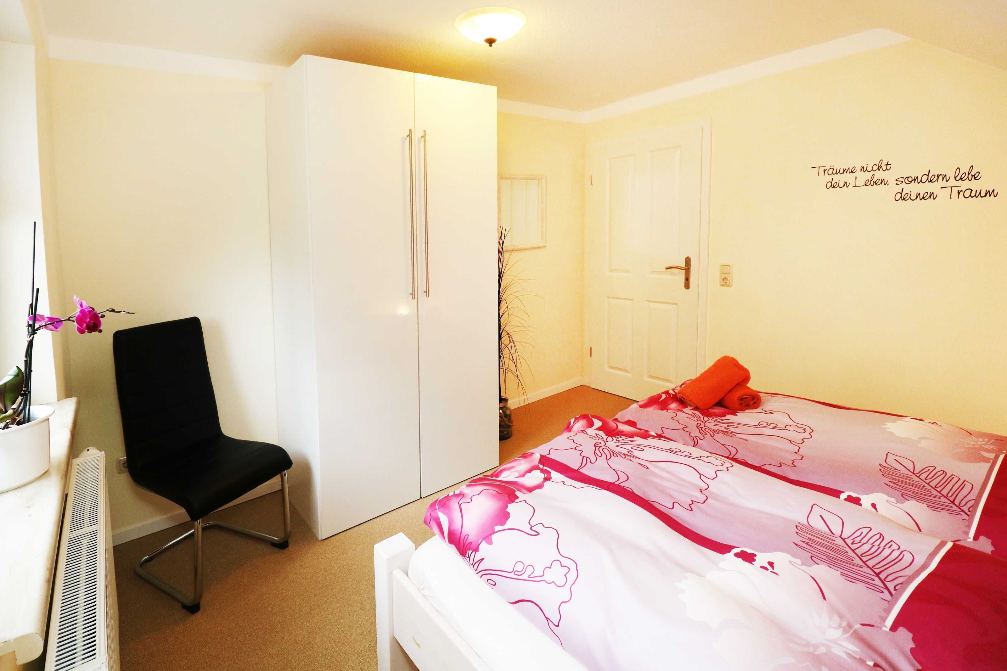 A bedroom with a bed, a chair, a wardrobe, and a wall with a motivational quote.
