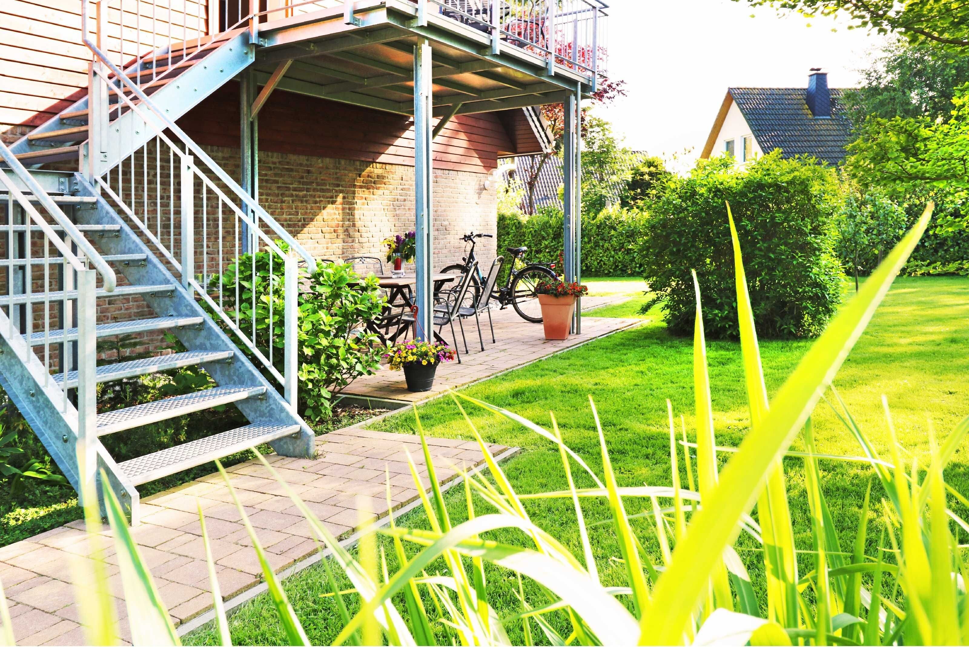 Ein Wohngebäude mit überdachter Veranda, einer Treppe und einem Garten mit Rasen und Pflanzen.