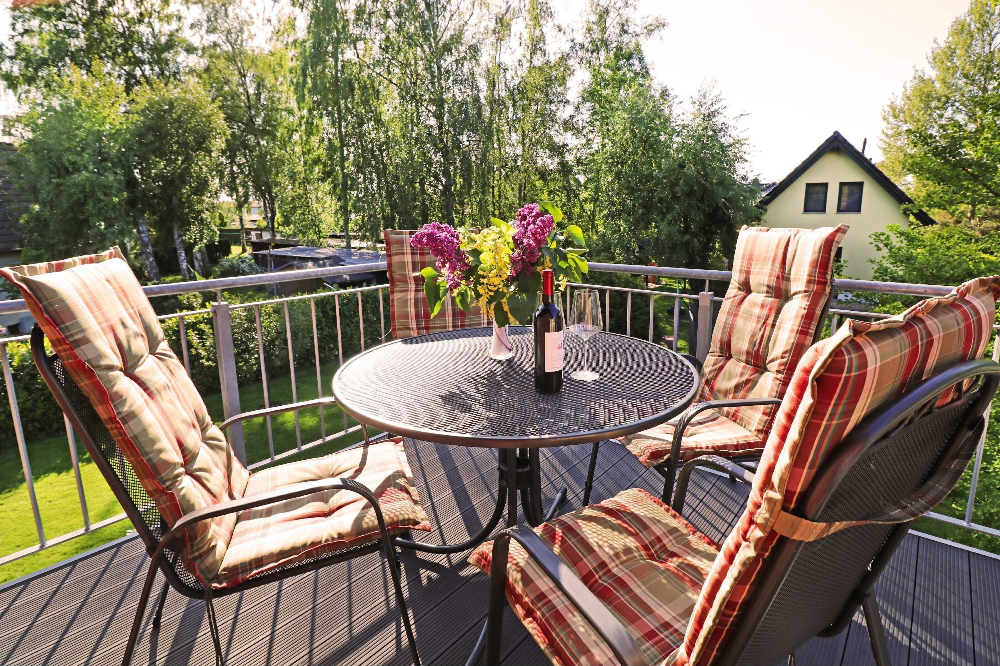 A round metal table with two chairs, each with plaid cushions, on a deck with a railing. A vase with flowers and a bottle of wine are on the table.