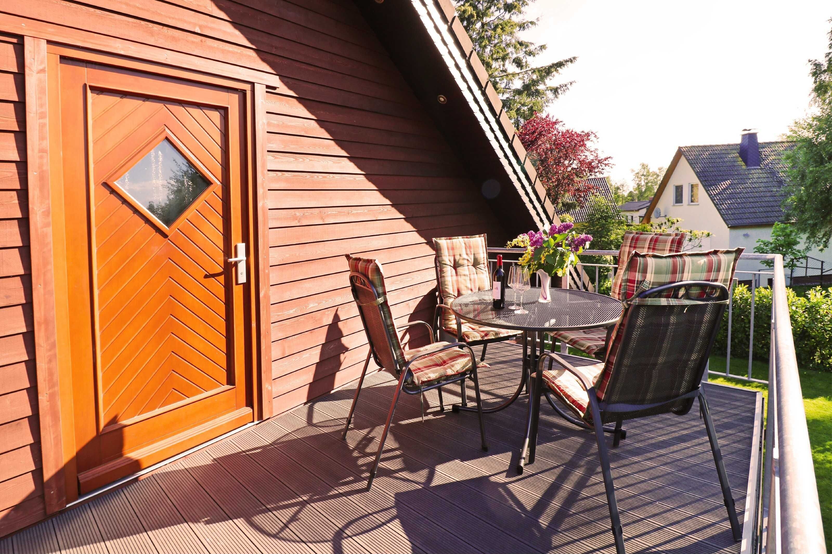 Eine Holzterrasse mit Tisch und Stühlen, eine Tür mit einem rautenförmigen Fenster und ein Blick auf ein Haus im Hintergrund.