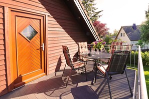 Una terraza de madera con una mesa y sillas, una puerta con un ventanal en forma de diamante, y vista a una casa al fondo.