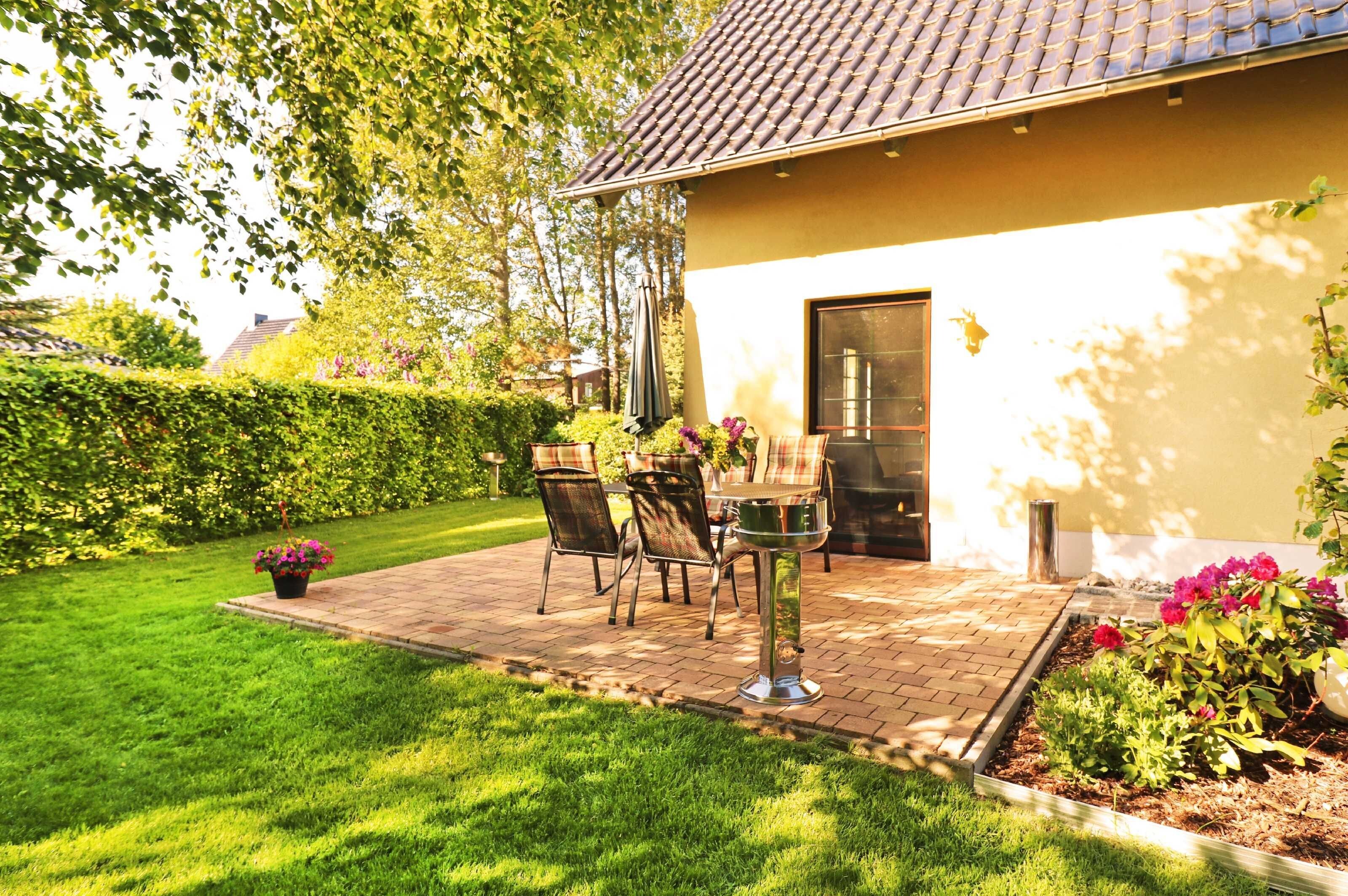 Eine Terrasse mit Tisch und Stühlen, umgeben von einem gepflegten Rasen und Blumenbeeten.