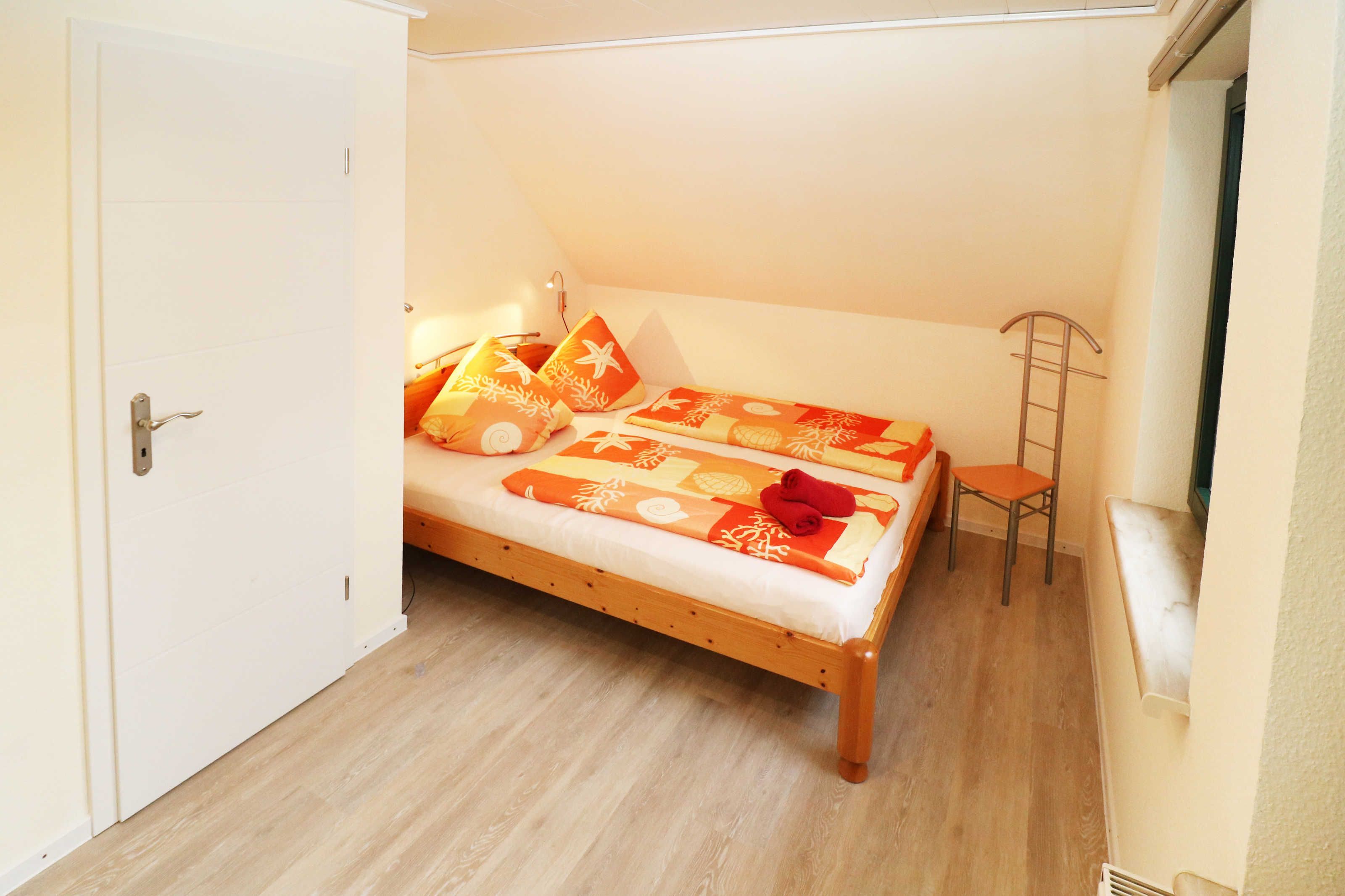 Ein kleines Zimmer mit einem Einzelbett, Holzboden, einer weißen Tür und einem kleinen Stuhl.