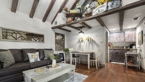 A cozy living room with a dark leather sofa, a white coffee table, and a dining area with a white table and chairs. A kitchen with wooden cabinets and a hanging light fixture is visible in the background. - Wildflower Cottage/PERFECT LOCATION!!/Walk to Main/Marktplatz!! (Fredericksburg)