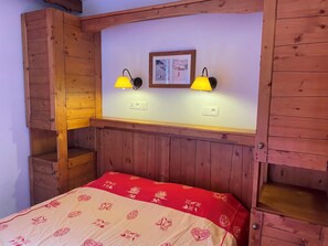 A wooden bunk bed with two bedside lamps, a framed picture on the wall, and a red and beige bedspread.