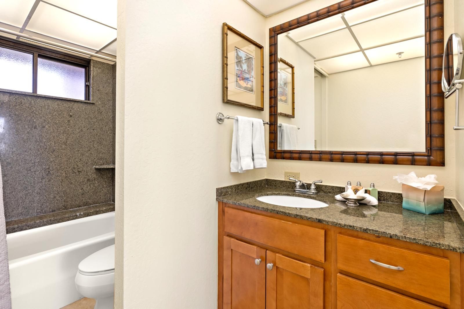 A bathroom with a granite countertop sink, wooden cabinets, a large mirror, and a bathtub.