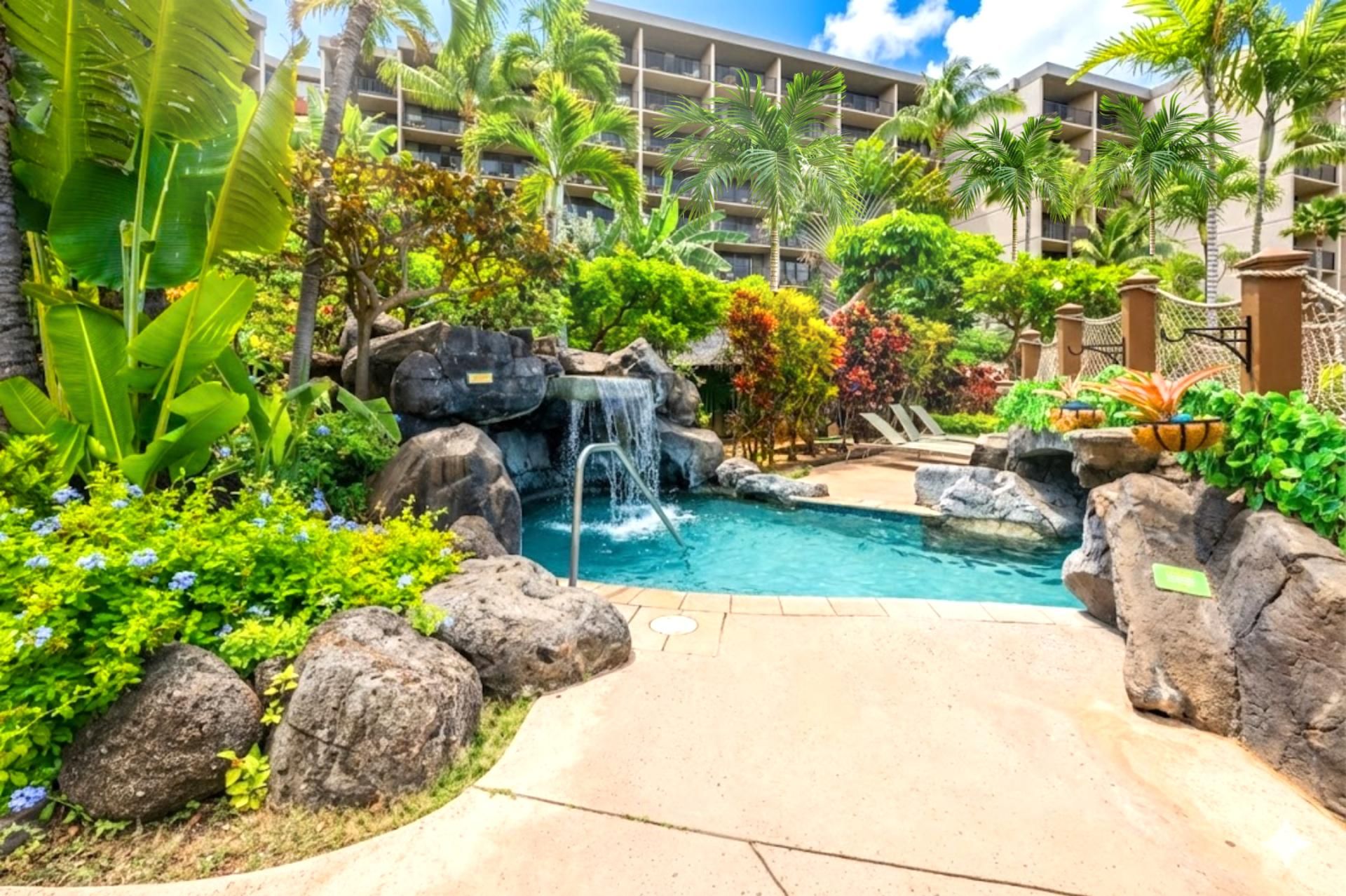 A pool area with a waterfall, surrounded by tropical plants and palm trees, with a building in the background.