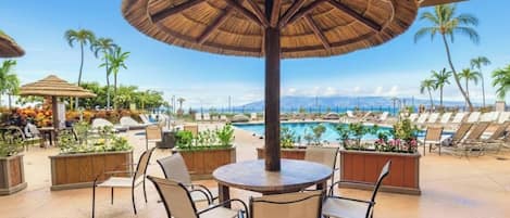 A poolside area with a round table and chairs, a thatched umbrella, and lounge chairs.