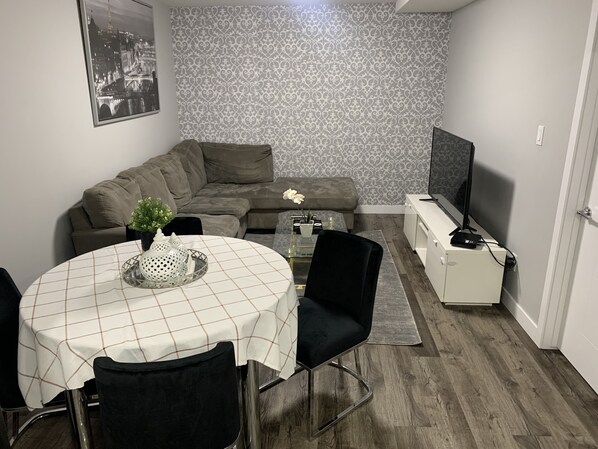 A living room with a sofa, a dining table with chairs, a TV, and a patterned wallpaper.
