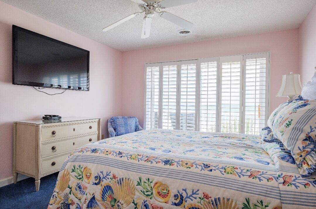 A bedroom with a bed, a television mounted on the wall, a ceiling fan, and a window with blinds.