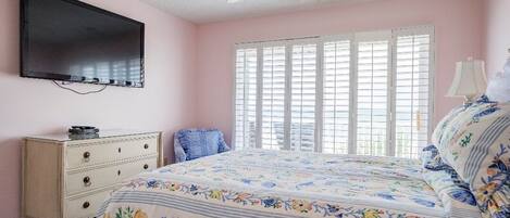 A bedroom with a bed, a television mounted on the wall, a ceiling fan, and a window with blinds.