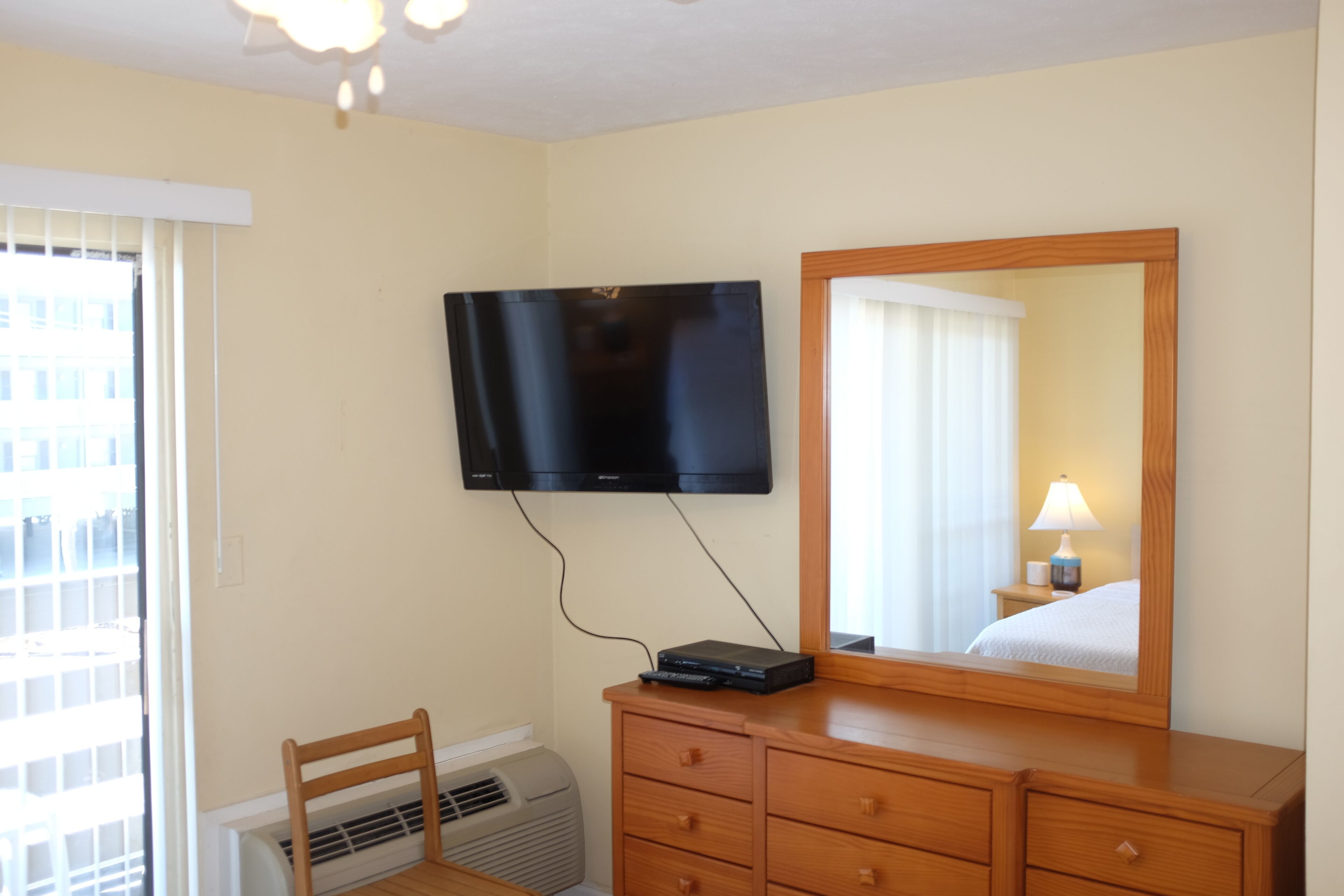 Une chambre d’hôtel dotée d’un téléviseur à écran plat fixé au mur, d’une commode en bois, d’un miroir et d’une fenêtre avec des stores.