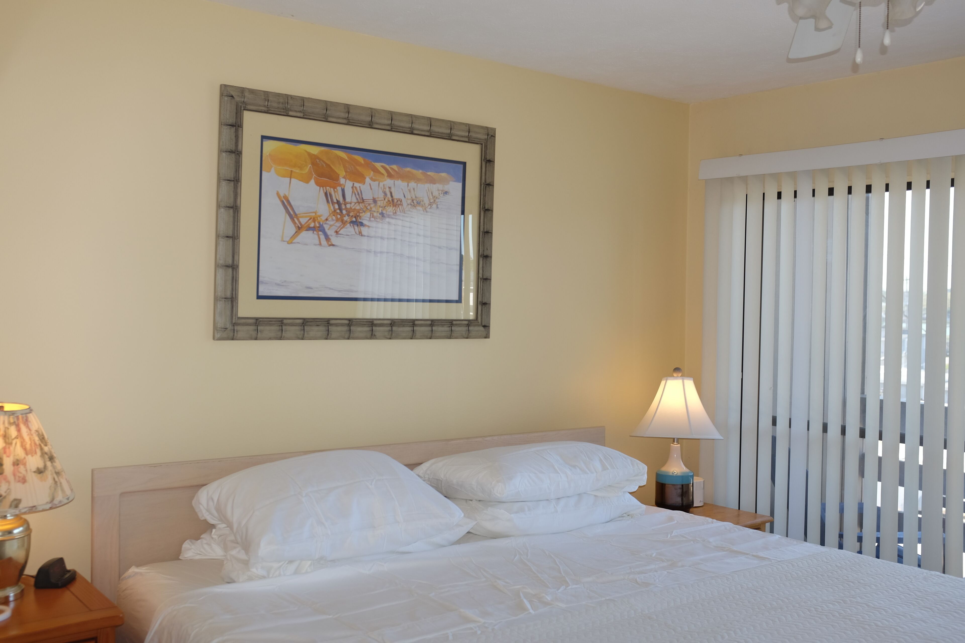 Een hotelkamer met een bed, een nachtkastje, een lamp, een lijst met een foto aan de muur en een raam met jaloezieën.