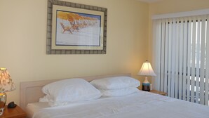 Et hotelværelse med en seng, et natbord, en lampe, et billede i ramme på væggen og et vindue med rullegardiner.
