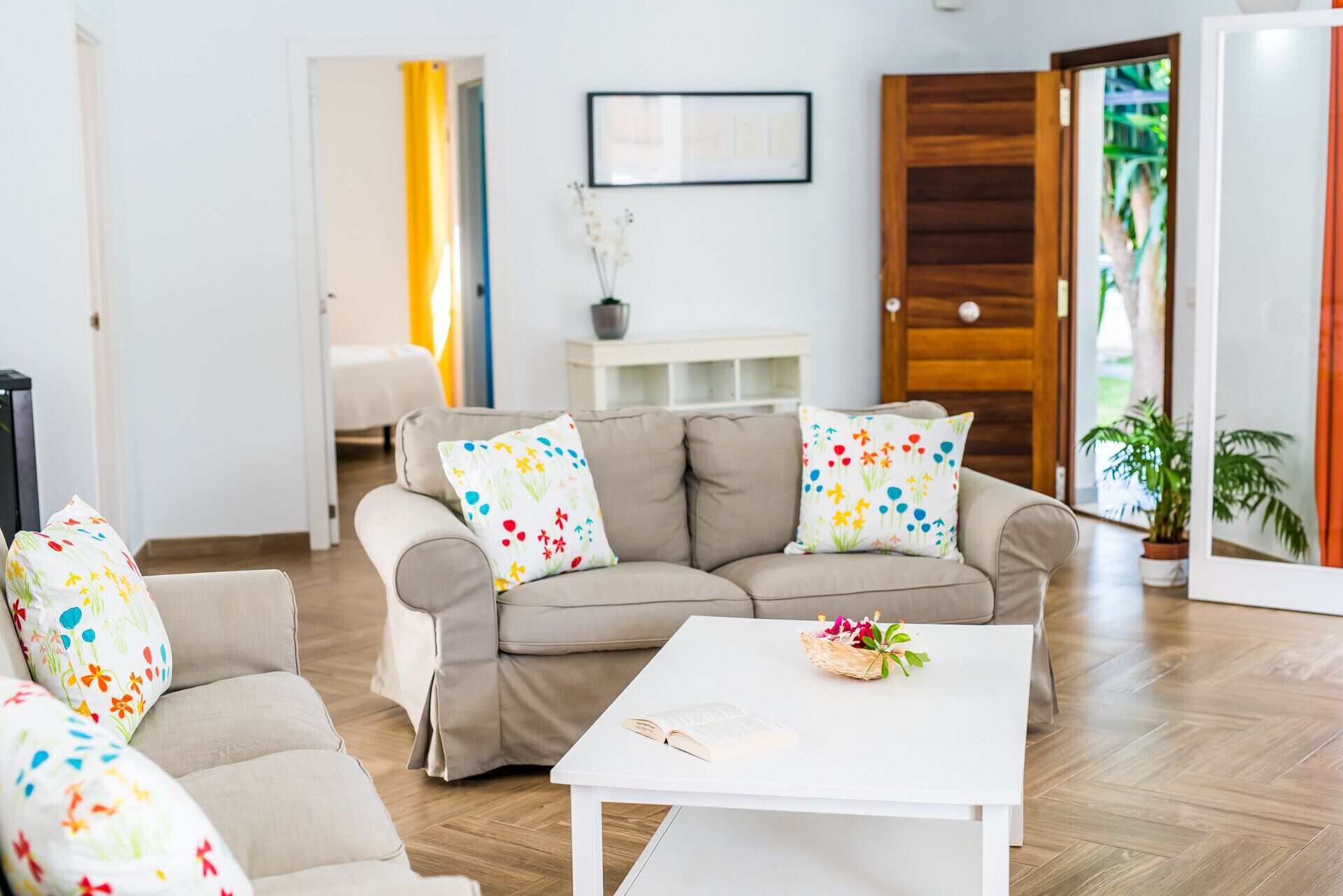 A living room with a beige sofa, colorful floral cushions, a white coffee table with a book and flowers, and a wooden door.