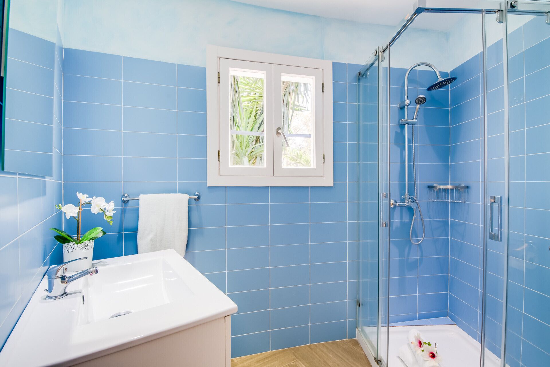 Een badkamer met blauwe tegels, een witte wastafel, een glazen douchecabine en een raam met uitzicht op groen.