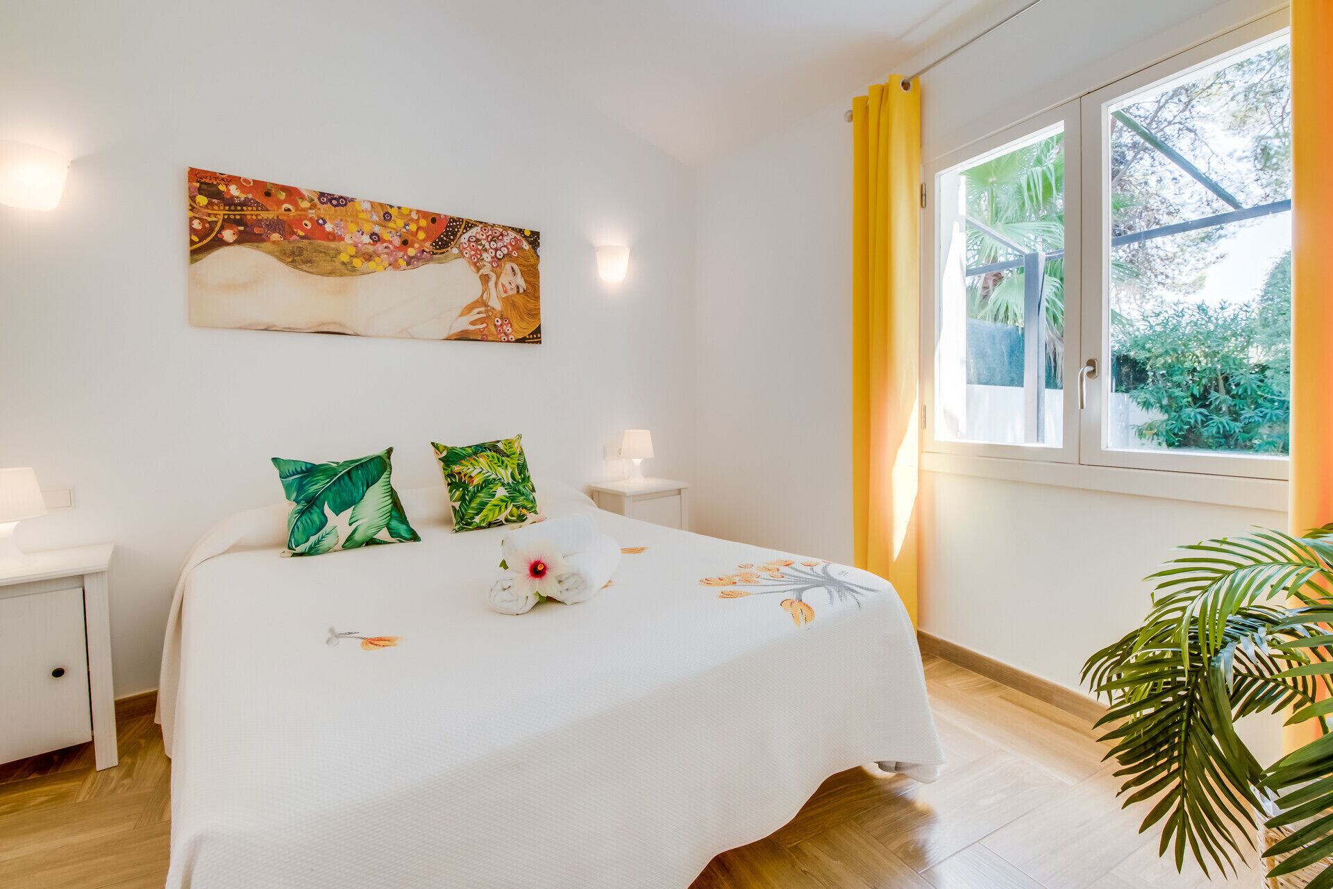 A bedroom with a bed, pillows, a nightstand, a lamp, a window with curtains, a plant, and a painting on the wall.