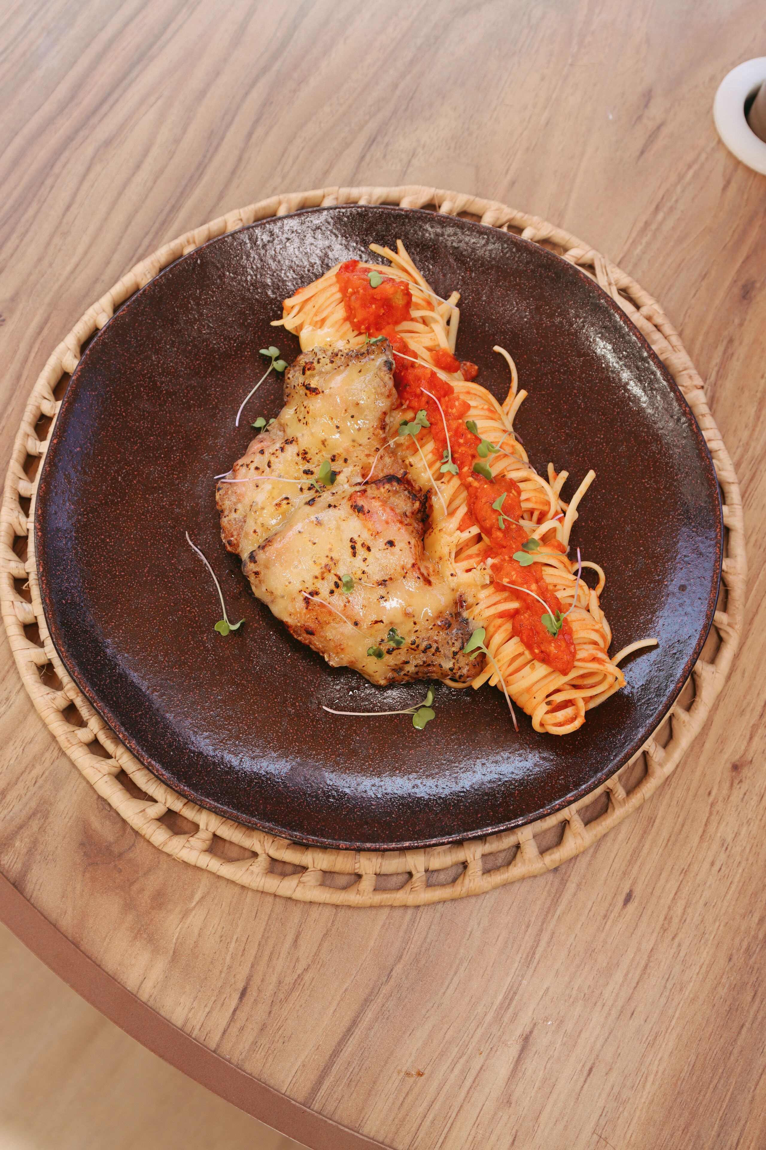 A plate of spaghetti with a piece of meat and tomato sauce, garnished with herbs, on a dark ceramic plate.