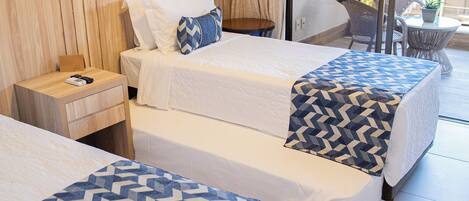 A hotel room with two beds, a wooden headboard, a small table with a chair, and a view of the outside balcony.