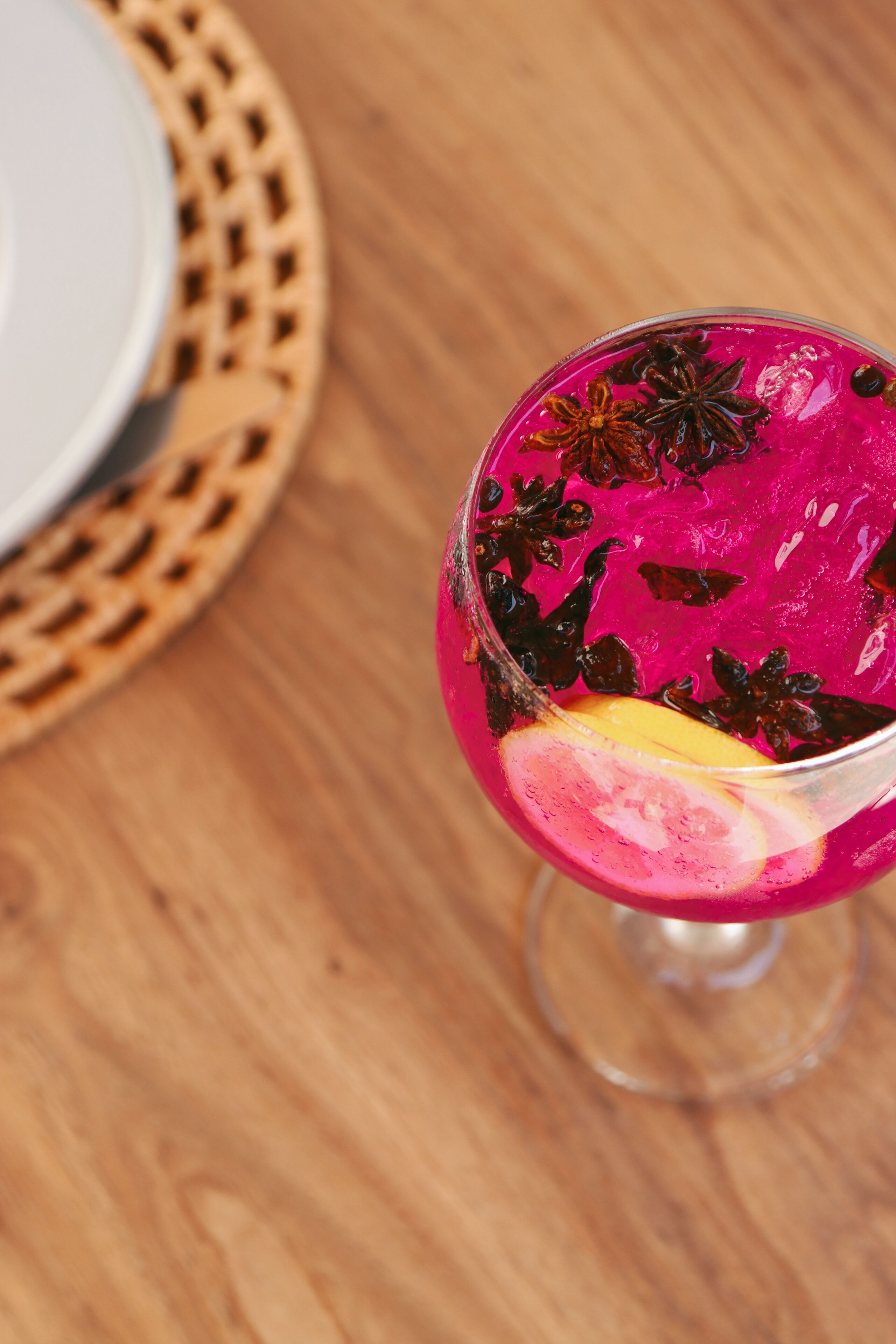 Un vaso de cóctel con una bebida rosa brillante, adornado con anís estrellado y una rodaja de limón, sobre una mesa de madera.