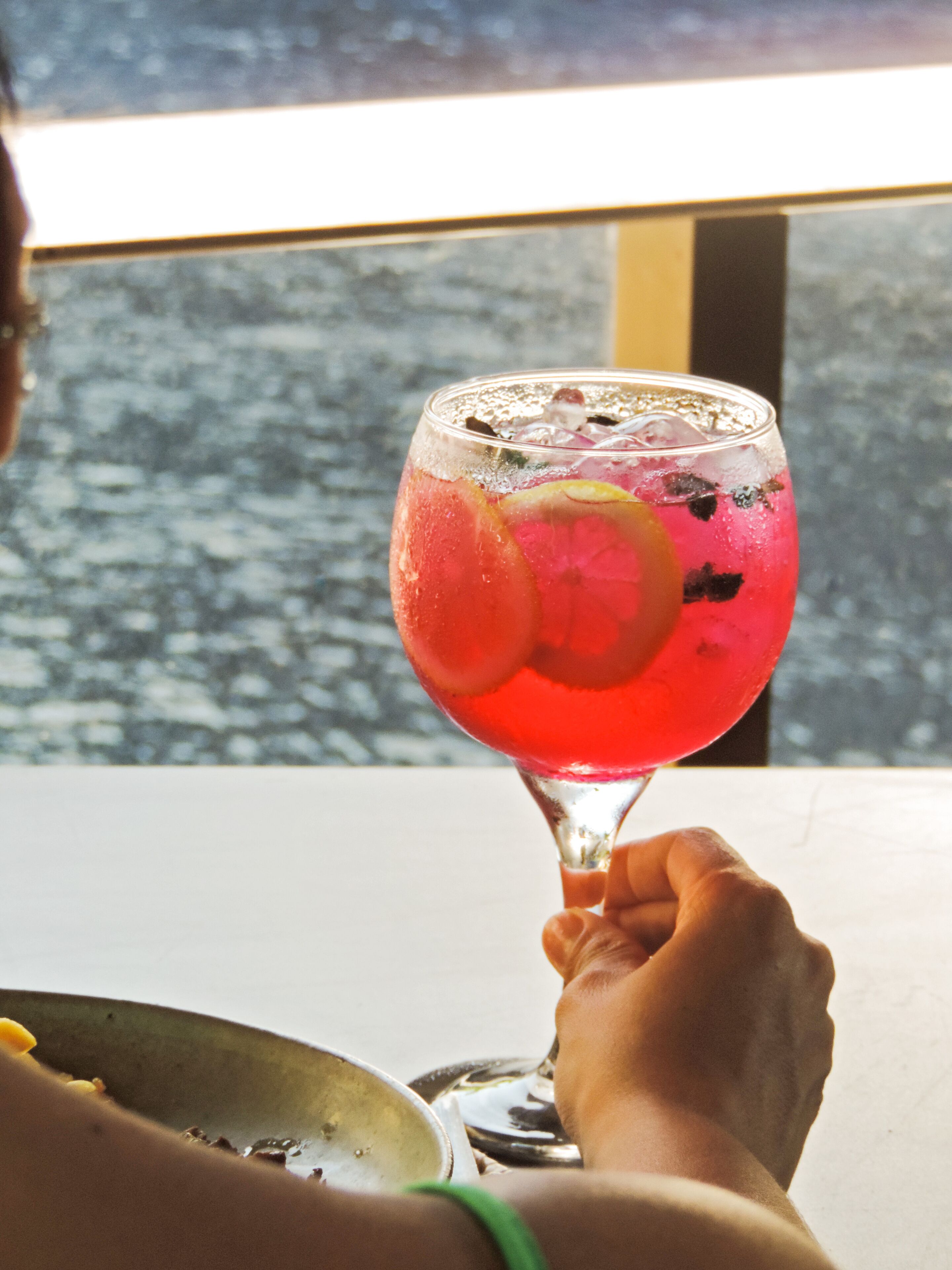 A person holding a glass with a red drink, a green bracelet, and a metal bowl on a table.