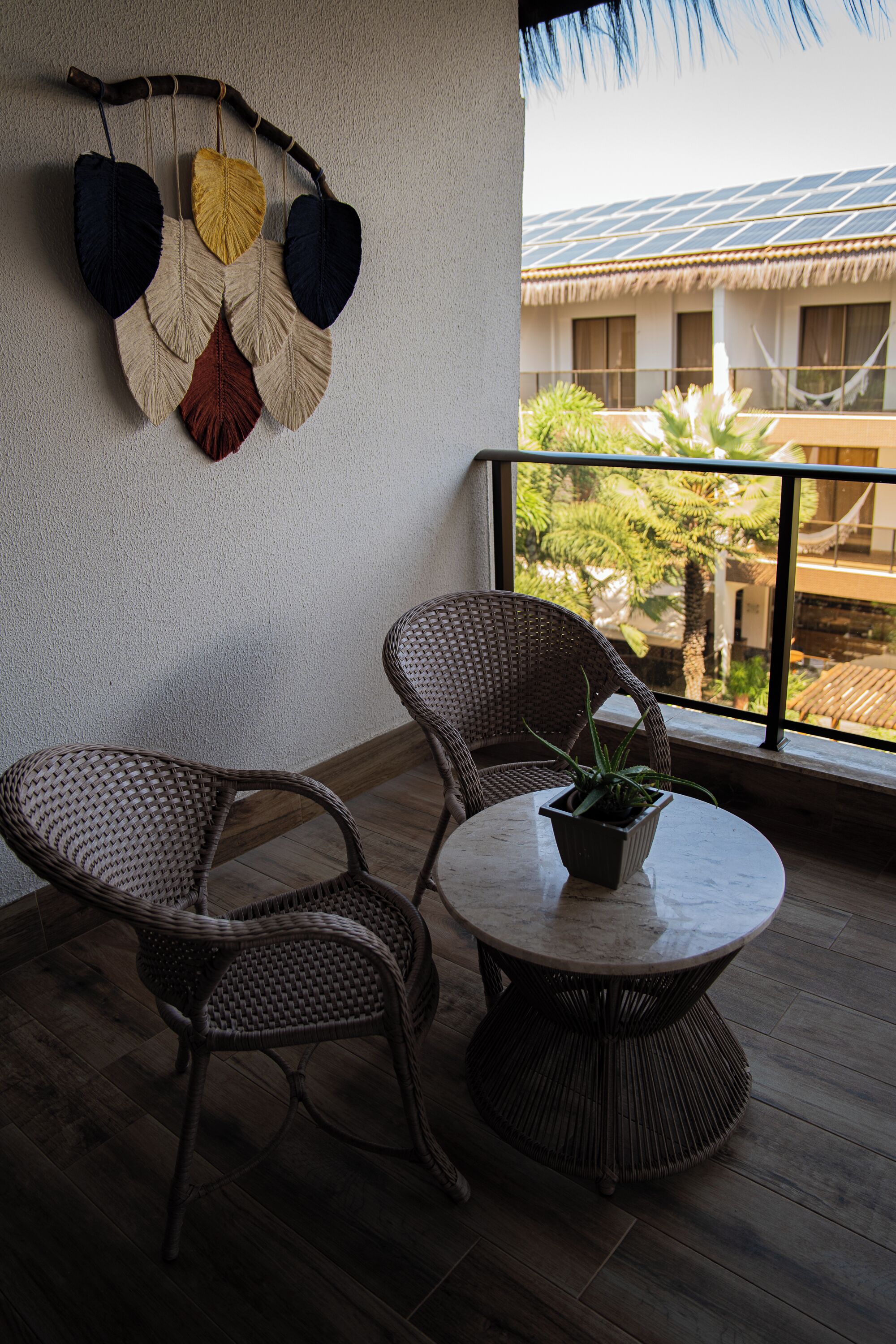 A balcony with wicker chairs and a round table, a wall hanging with leaves, and a view of a building with a tiled roof.