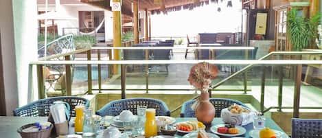 A dining area with tables set for breakfast, wicker chairs, and a thatched roof.