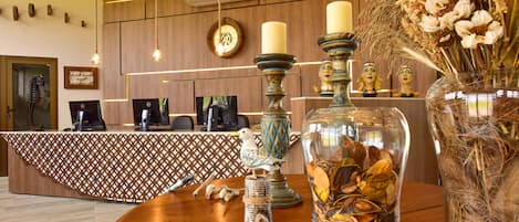 Un mostrador de recepción con mantel estampado, mostrador de madera, un reloj y objetos decorativos, incluyendo una figurina de pájaro, velas y un jarrón con flores secas.