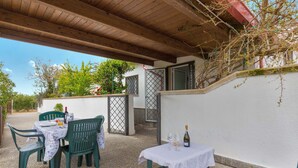 Un patio al aire libre con techo de madera, sillas de plástico verde, una mesa con mantel blanco y una botella de vino.