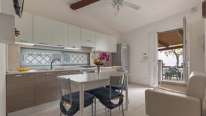 Una cocina moderna con armarios blancos, una mesa de comedor y sillas. Hay un ventilador de techo, una ventana con persianas y vistas al exterior.