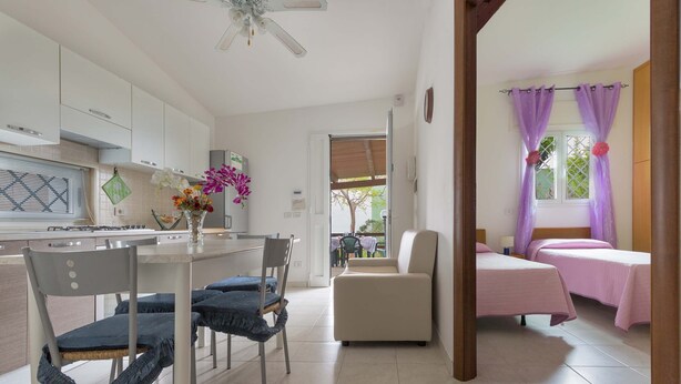 Una cocina moderna con armarios blancos, una mesa de comedor con sillas y un florero con flores. Hay dos camas con ropa de cama rosa y cortinas moradas.