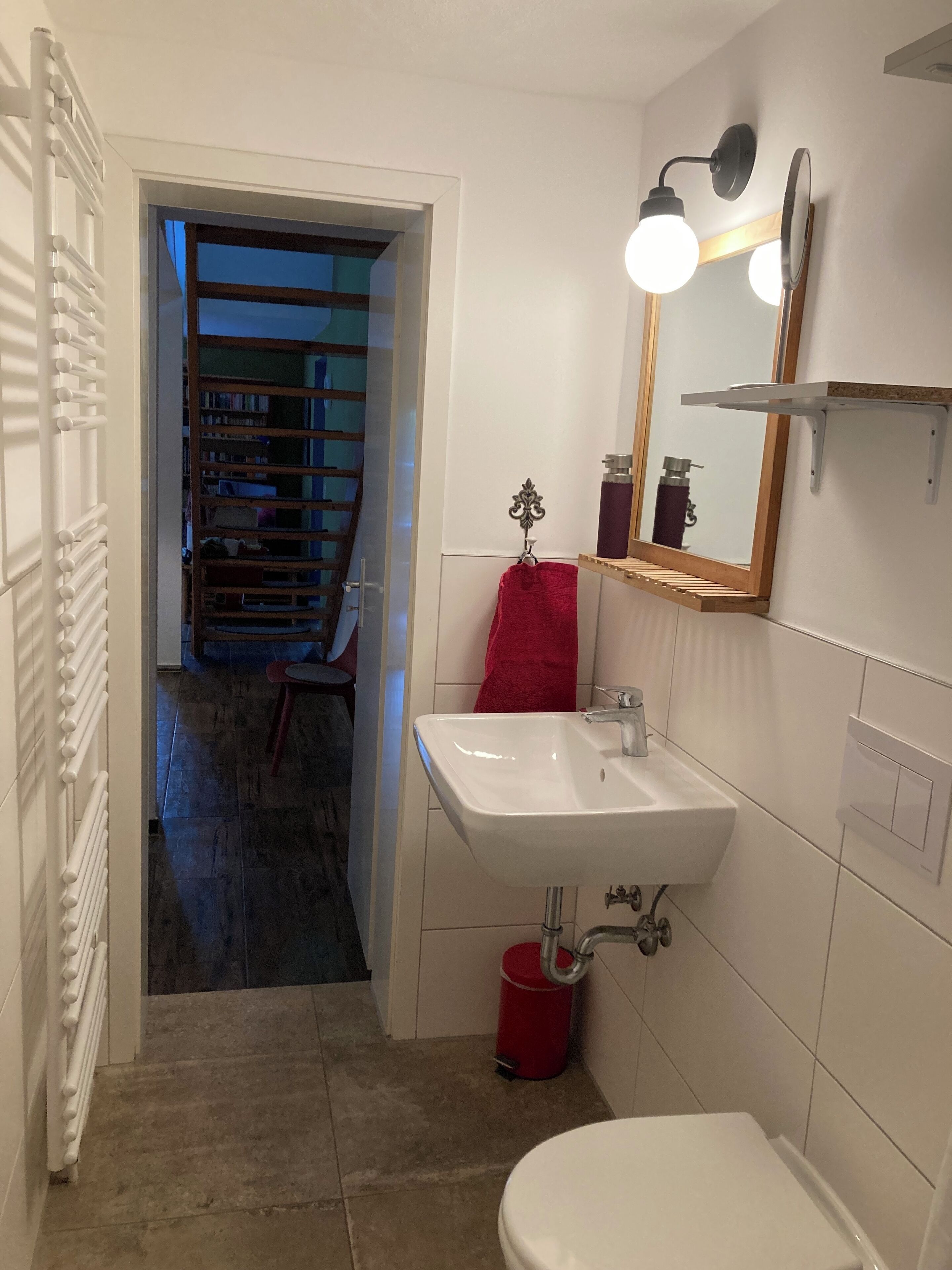 Une salle de bain avec un lavabo blanc, un miroir avec éclairage, une serviette rouge et une poubelle rouge.