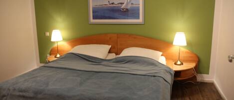 Un lit bien fait, recouvert d’une couverture bleue, avec deux lampes de chevet et un tableau représentant un voilier accroché au mur.