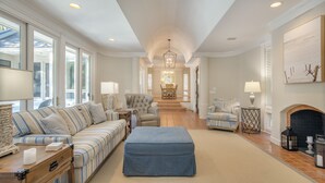 A spacious living room with a sofa, ottoman, and armchairs, a fireplace, and a dining area in the background.