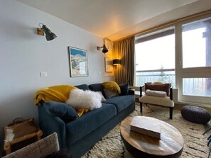 A living room with a dark blue sofa, a wooden coffee table, a chair, and a large window with curtains.
