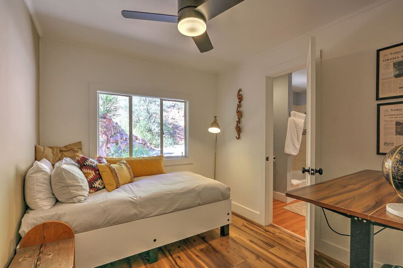 Un dormitorio con cama, ventilador de techo, ventana, lámpara de noche y escritorio de madera.