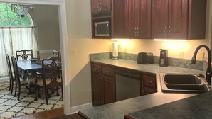 Una cocina con armarios de madera oscura, microondas, lavavajillas y un fregadero. Hay una mesa de comedor con sillas y una araña.